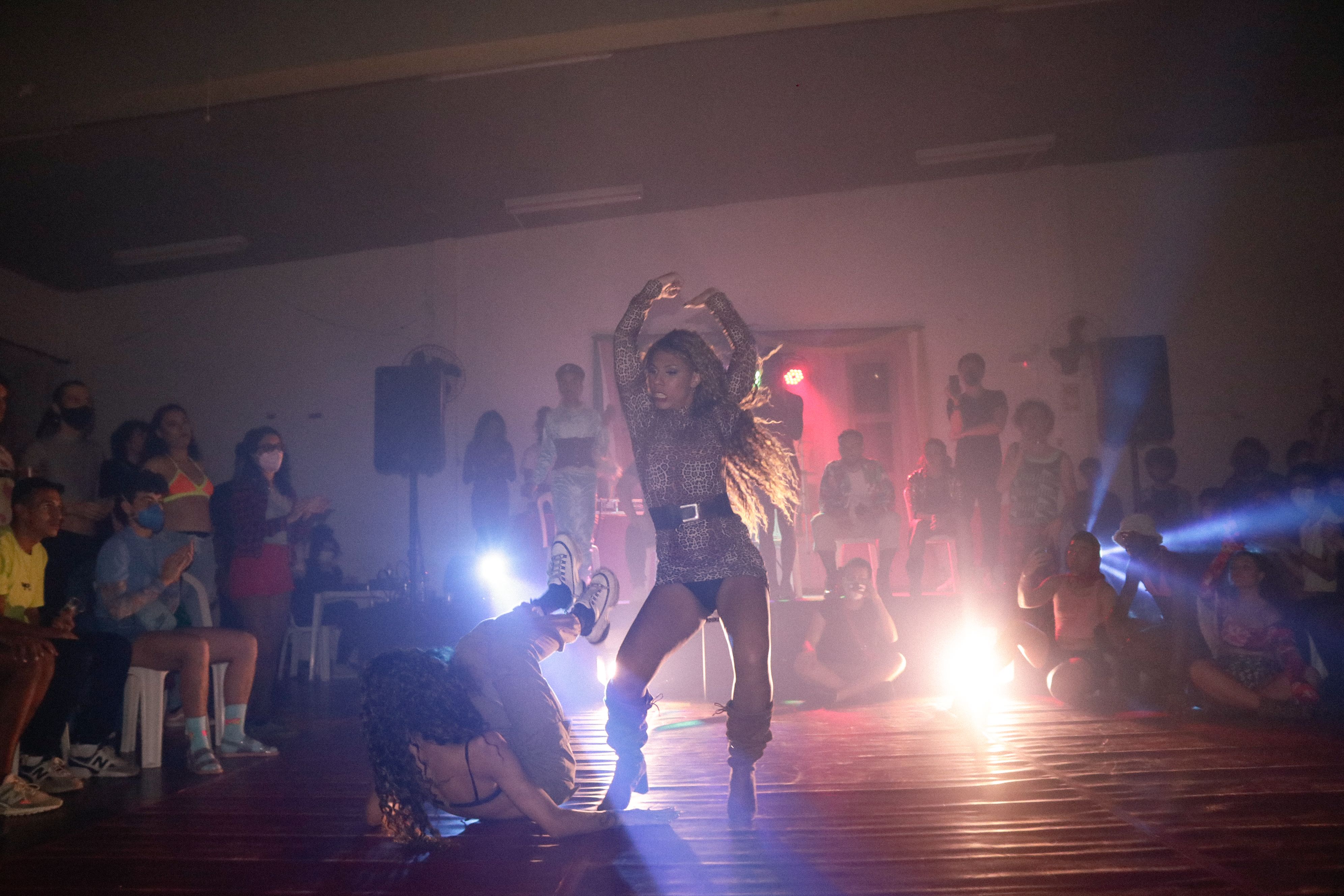 Pessoas dançando em frente a um público em sala iluminada com holofotes coloridos.