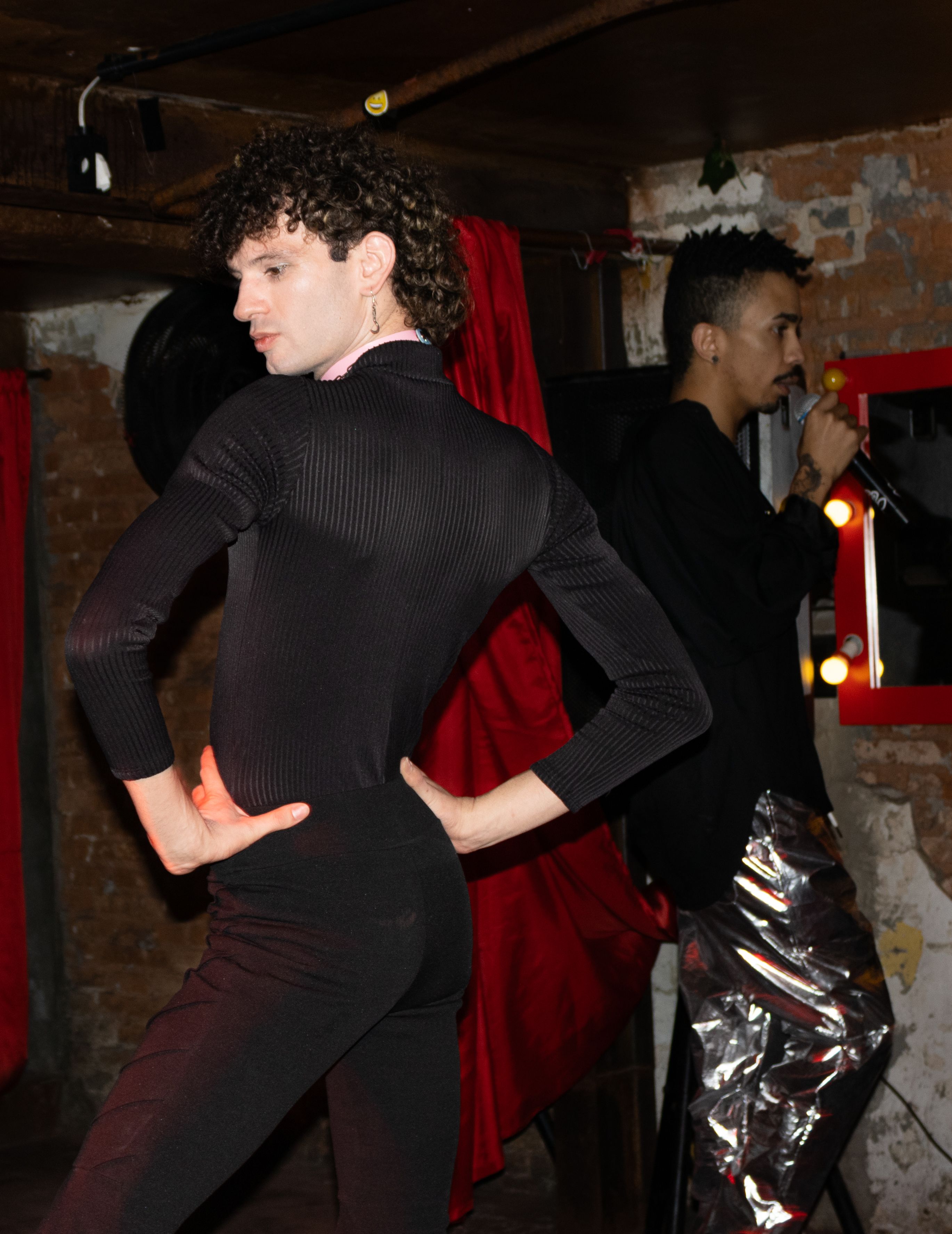 Duas pessoas, uma dançando em primeiro plano e outra em segundo plano com um microfone.